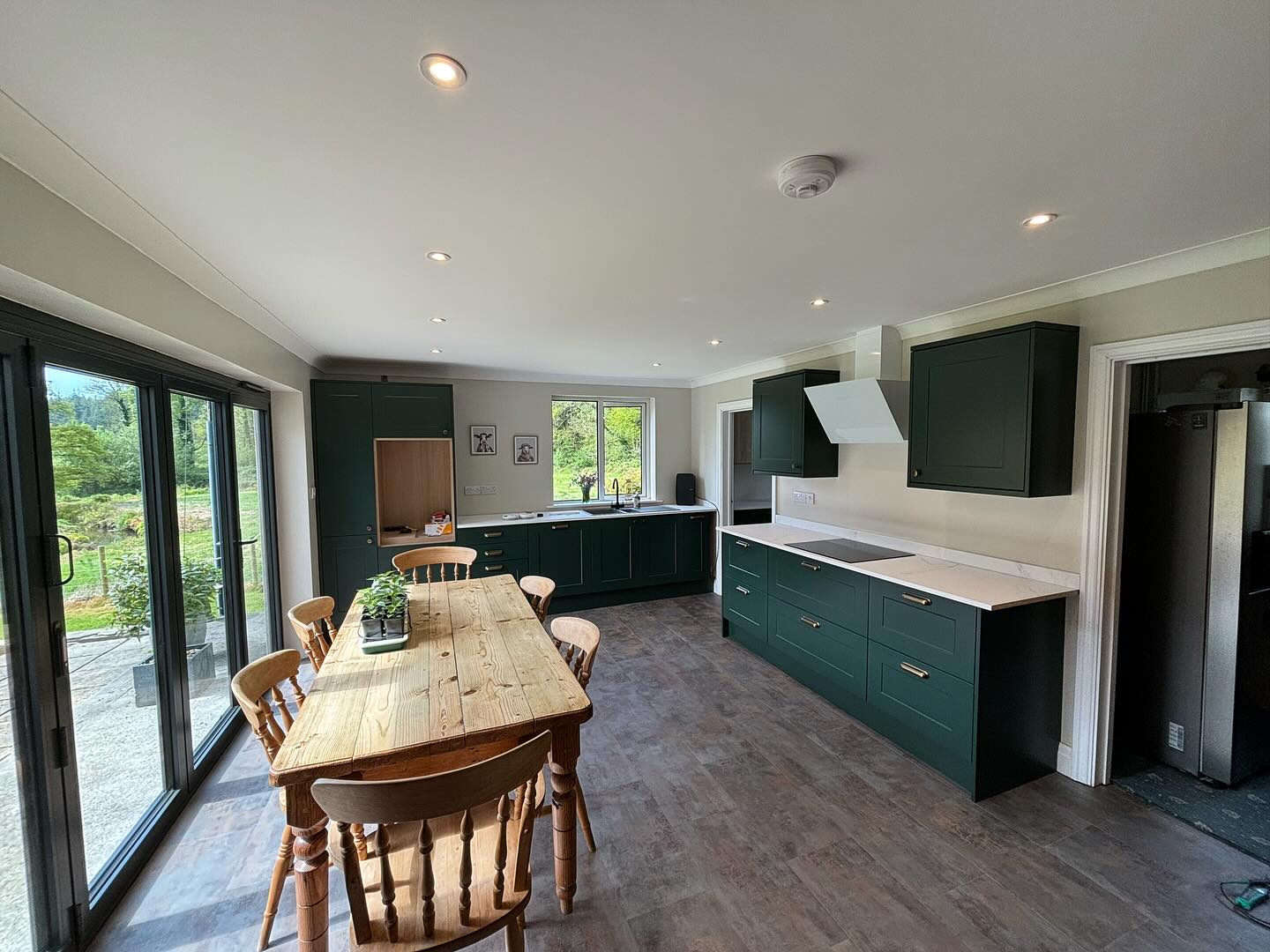 Finished green kitchen with down lights over a long wooden dining table and bifold doors