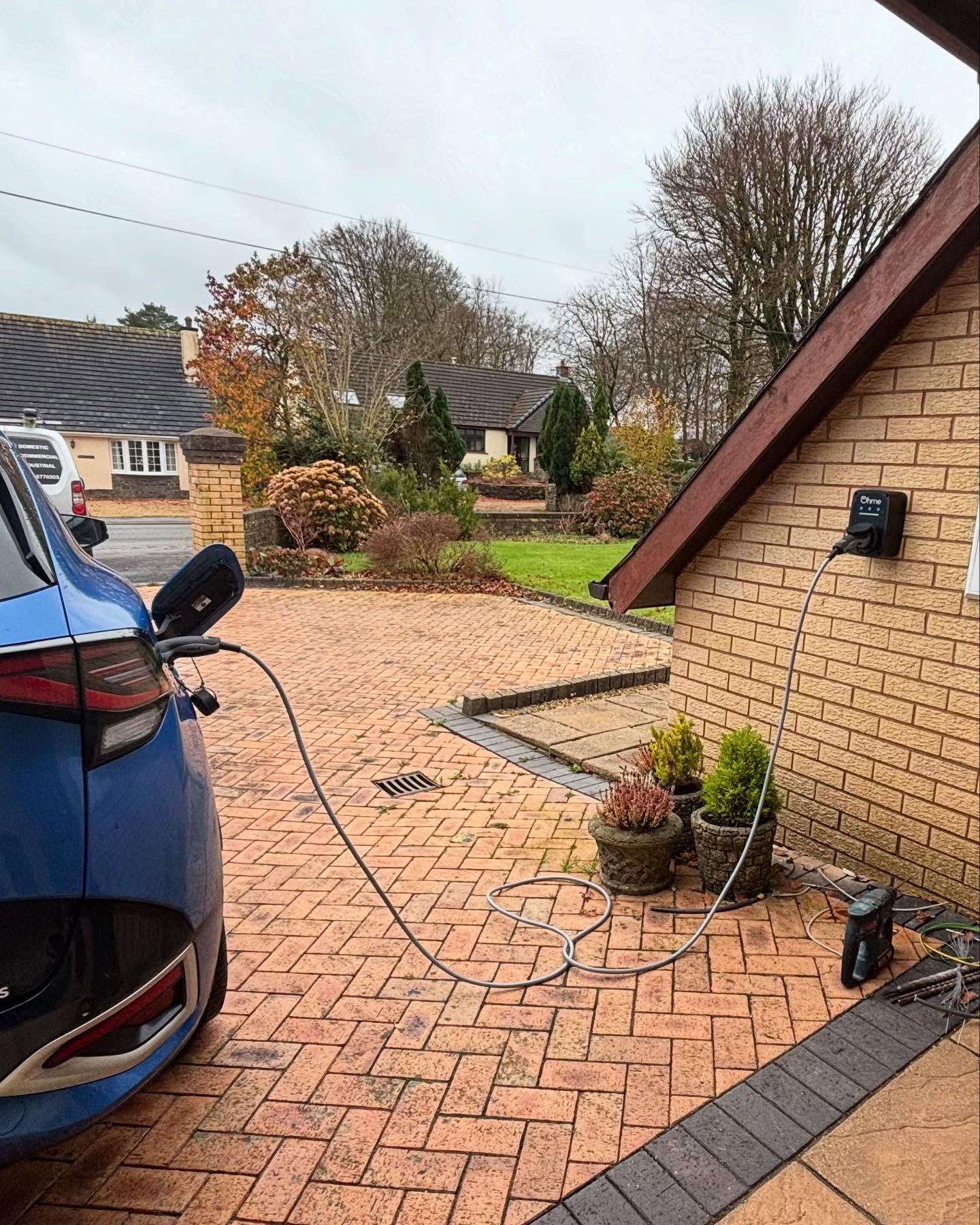 Blue electric car charging on a block-paved drive, with the TLEC van parked on the road behind